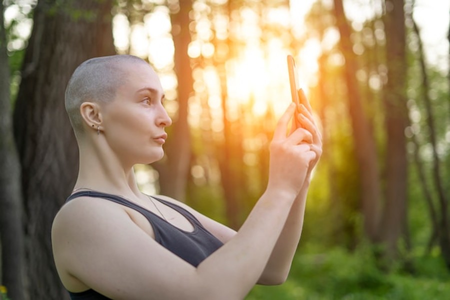 Une femme au crâne rasé utilise son smartphone pour suivre sa routine de biohacking wellness au cœur d'une forêt lumineuse.