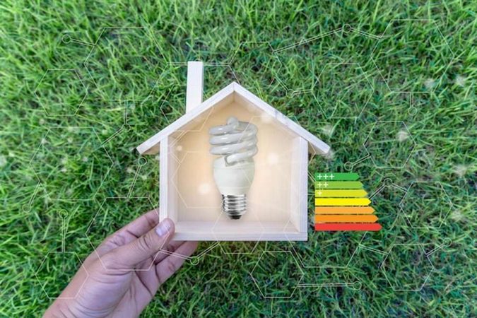 Une main tenant une petite maison en bois avec une ampoule basse consommation pour découvrir comment domotiser son tableau électrique pour réduire sa facture d'énergie : Le guide complet.