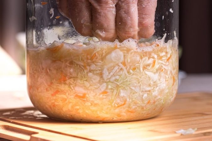 Une main presse fermement des légumes râpés dans un bocal en verre pour extraire leur jus naturel, illustrant les principes fondamentaux du guide 'Comment fabriquer son ail noir maison sans machine : Le guide complet de la fermentation lente'.