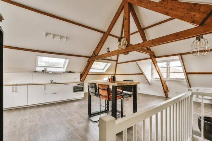 Un grenier mansardé sert actuellement de zone de stockage avec des malles anciennes et une planche à repasser. Cet espace étroit aux poutres apparentes blanches montre le potentiel de rénovation pour ceux qui souhaitent savoir comment transformer une charpente fermette en combles aménageables : étapes, prix et autorisations sont nécessaires pour convertir ce volume en une pièce de vie confortable et lumineuse.