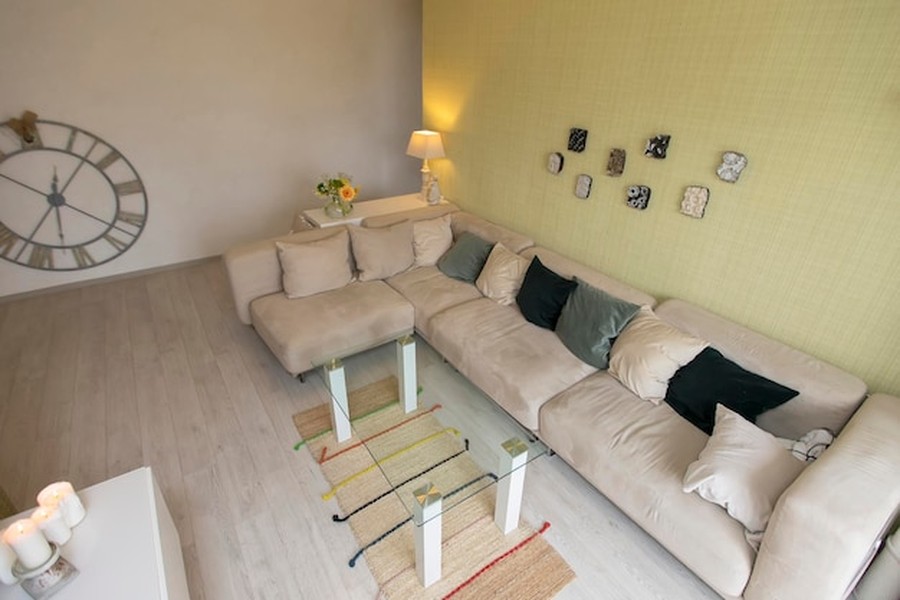 Canapé d'angle beige clair dans un petit salon aménagé, avec table basse en verre et grande horloge murale décorative.