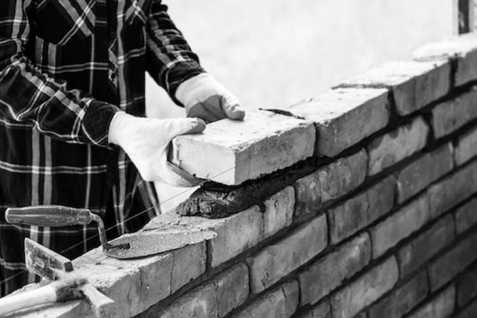 Un artisan maçon pose avec précision une brique sur un lit de mortier, illustrant la Maçonnerie et RGA : Les nouvelles techniques de fondations pour sols argileux instables en 2026.