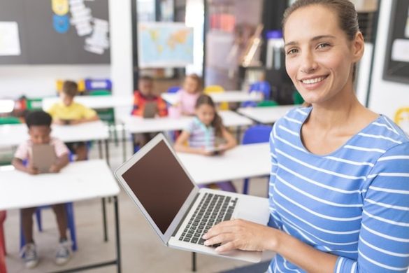 Une enseignante souriante utilise son ordinateur portable dans une classe numérique, illustrant la Reconversion : Comment devenir Concepteur Pédagogique quand on est enseignant (Guide complet).