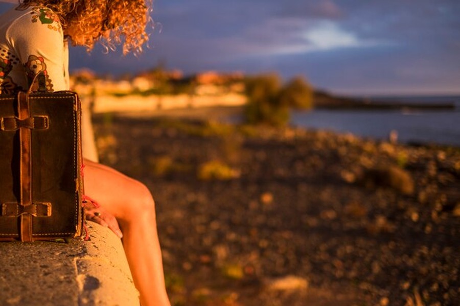 Une voyageuse contemplative assise au bord de l'océan avec sa valise vintage pour un séjour en mode Cool-cationing : les meilleures destinations pour fuir la chaleur sans renoncer aux paysages de rêve.
