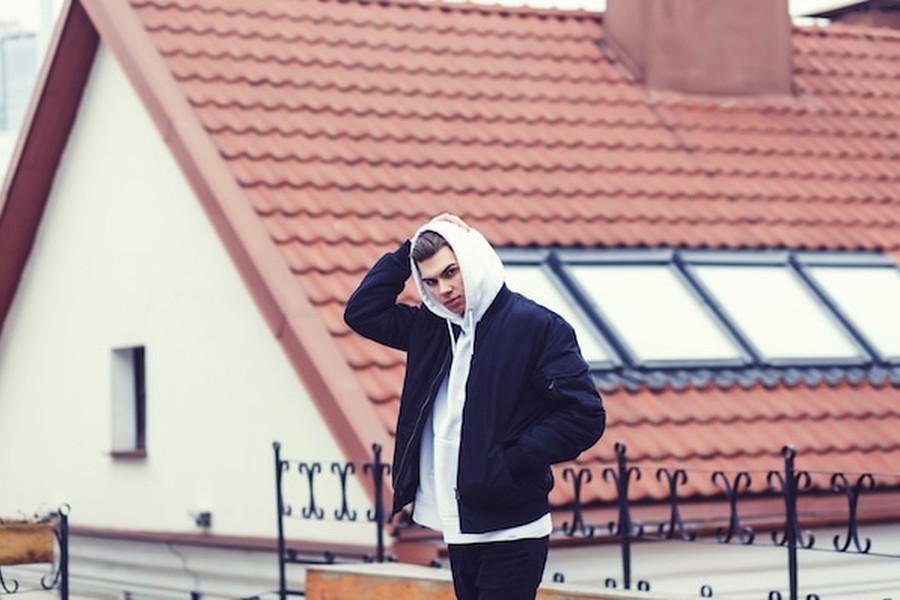 Un jeune homme pensif devant une maison à toit de tuiles traditionnelles, illustrant pourquoi votre assurance toiture va changer en 2026 face aux risques de grêle.