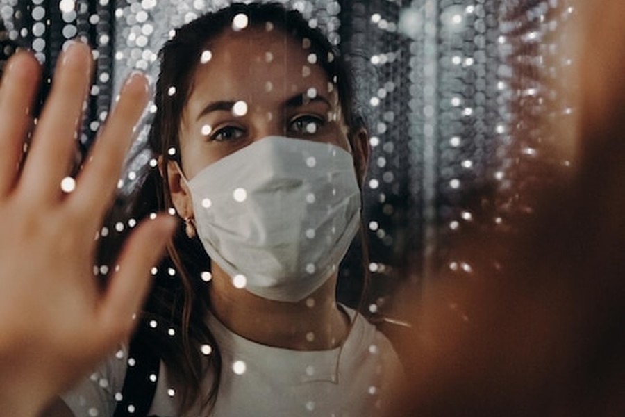 Une jeune femme portant un masque de protection regarde à travers une vitre parsemée de reflets lumineux, illustrant comment protéger son identité numérique contre le clonage vocal et visuel.