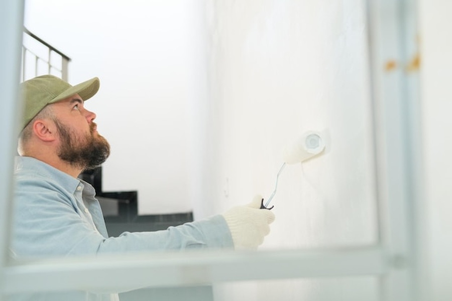 Un homme barbu en casquette applique soigneusement une Peinture thermorégulante intérieure : le guide complet pour réduire la chaleur de ses murs sans climatisation à l'aide d'un rouleau.