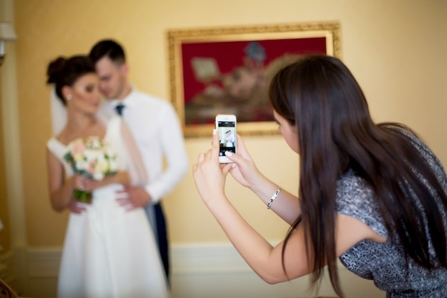 Une femme photographie un couple de jeunes mariés élégants, illustrant parfaitement pourquoi intégrer des hologrammes de proches disparus à sa cérémonie de mariage pour créer des souvenirs éternels.