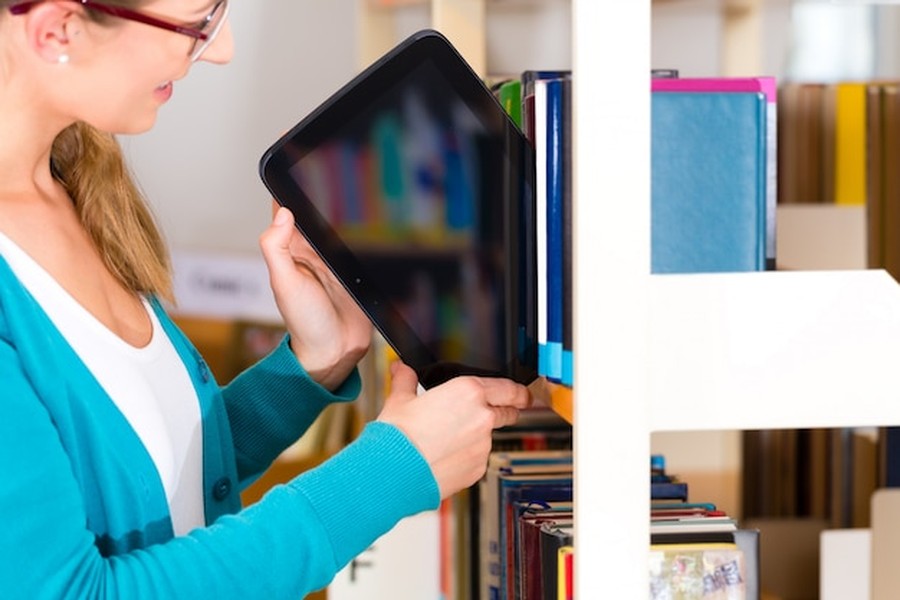 Une femme insère une tablette tactile entre des livres sur une étagère, illustrant comment bâtir sa propre bibliothèque numérique pérenne : le guide de l'archivage culturel personnel.