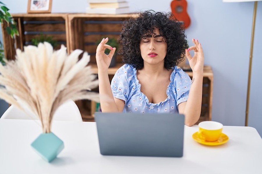 Une femme aux cheveux bouclés médite devant son ordinateur pour apprendre comment pratiquer l'hormèse pour transformer sa résistance au stress et à la fatigue.