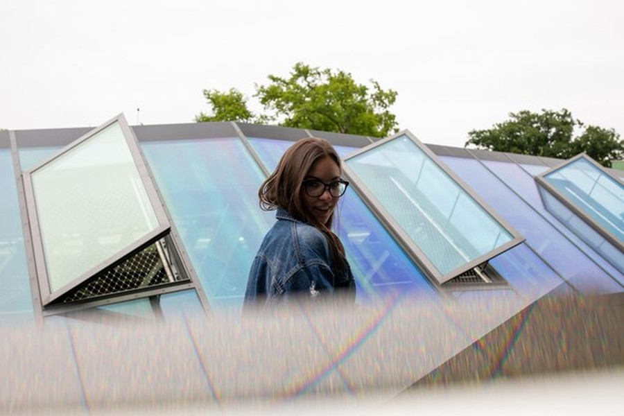 Une jeune femme se tient devant une toiture innovante en verre incliné, démontrant comment optimiser le rendement de ses vitrages photovoltaïques transparents : le guide complet.