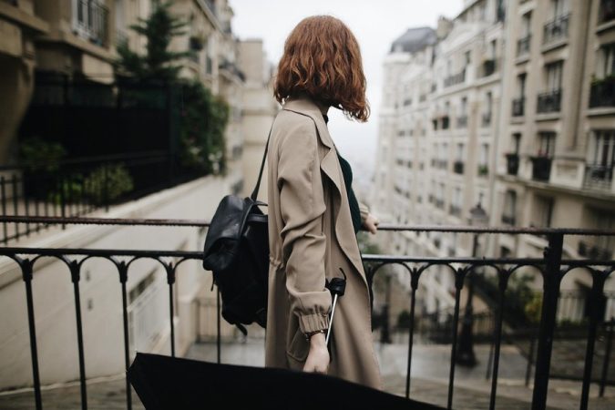 Une femme rousse en trench-coat admire l'architecture parisienne, cherchant de l'inspiration sur comment créer un potager d'intérieur automatisé avec des matériaux de récupération pour son appartement.
