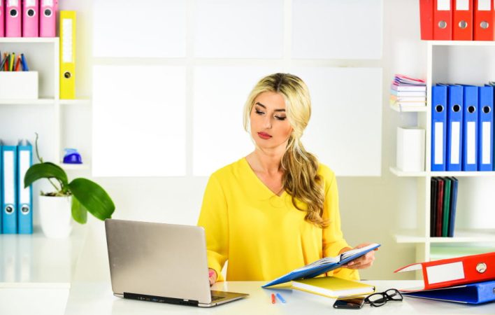 Une femme en haut jaune consulte un dossier devant son ordinateur pour télécharger un extrait kbis en ligne gratuit