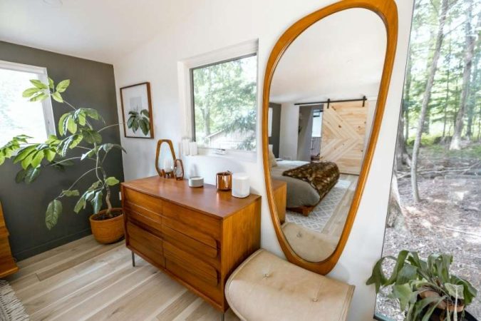 Un cluster de miroirs organiques et un grand miroir sur pied en bois encadrent une commode vintage dans une chambre lumineuse, montrant comment utiliser les miroirs pour décorer.