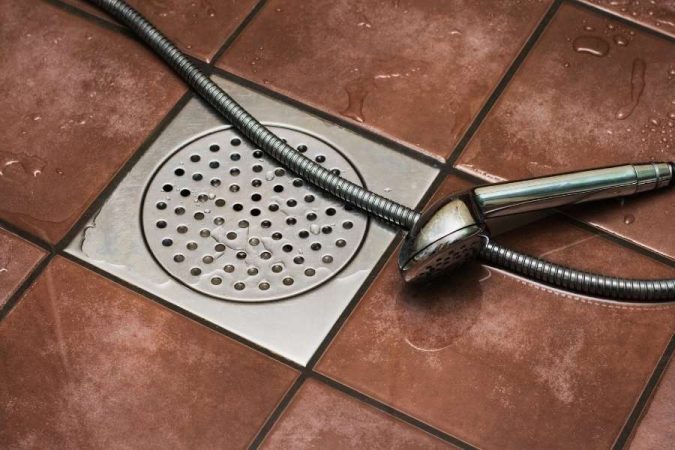 Un tuyau d'évacuation de douche et son pommeau chromés sur des carreaux de sol bruns.